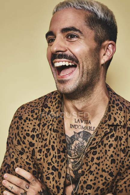 Portrait de gai modèle masculin à la mode avec des tatouages portant une chemise léopard à la mode debout sur fond beige et détournant les yeux — Photo de stock