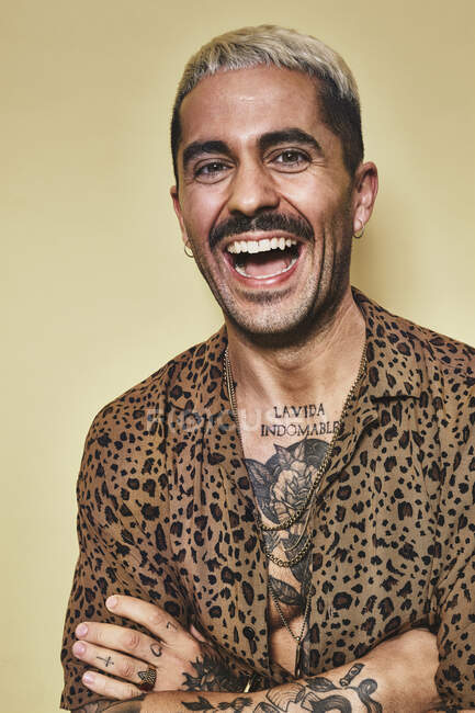 Retrato de alegre modelo masculino de moda con tatuajes con camisa de leopardo de moda de pie sobre fondo beige y mirando a la cámara - foto de stock