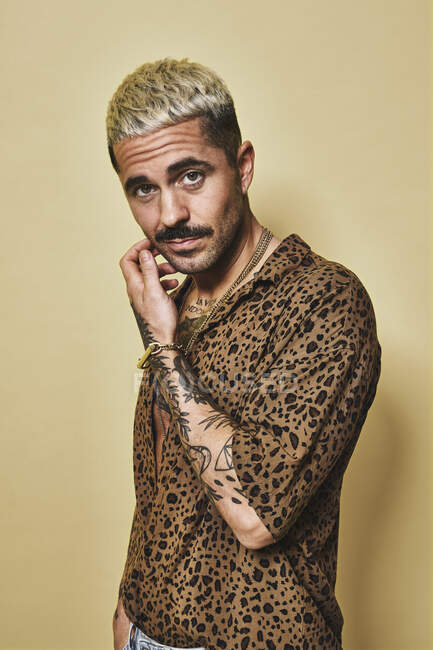 Modella maschile alla moda con tatuaggi che indossano camicia leopardata alla moda e jeans in piedi su sfondo beige e guardando la fotocamera — Foto stock