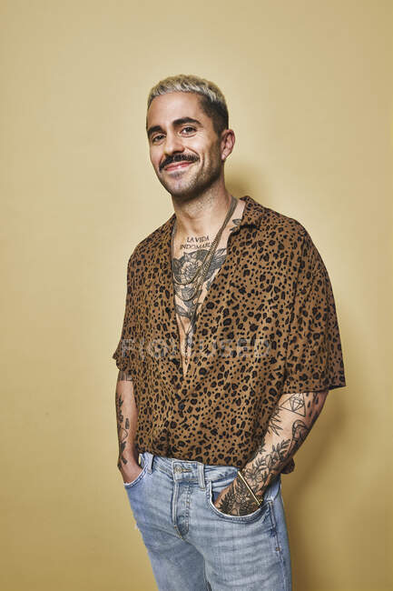 Модная мужская модель с татуировками в модной леопардовой рубашке и джинсах, стоящая на бежевом фоне и смотрящая в камеру — стоковое фото