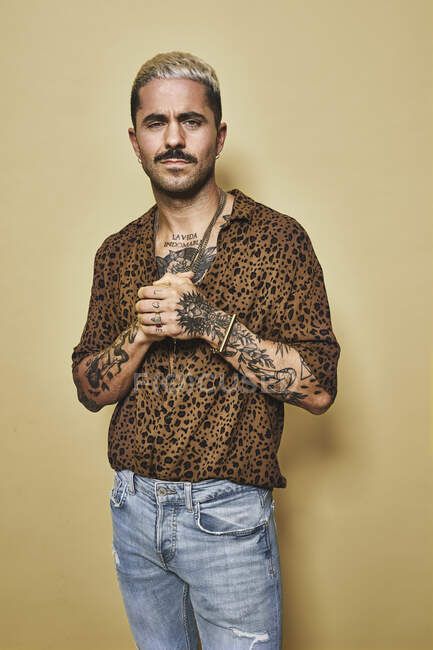 Modelo masculino de moda con tatuajes con camisa de leopardo de moda y jeans de pie sobre fondo beige y mirando a la cámara - foto de stock
