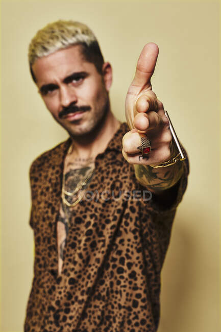 Adult bearded guy with stylish haircut and tattoo dressed in leopard shirt making finger gun gesture and looking at camera against yellow background — Stock Photo