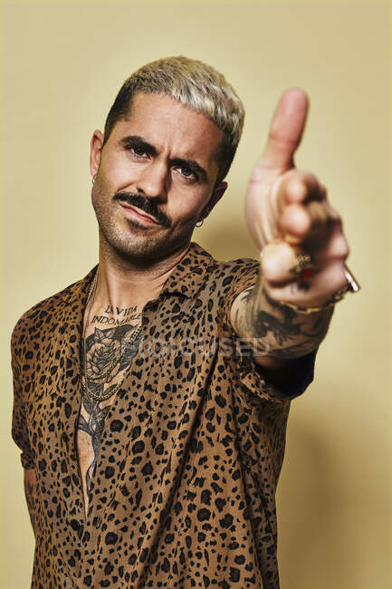 Взрослый бородатый парень со стильной стрижкой и татуировкой одетый в леопардовую рубашку делает жест пистолета и смотрит в камеру на желтом фоне — стоковое фото