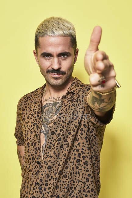 Hombre barbudo adulto con corte de pelo elegante y tatuaje vestido con camisa de leopardo haciendo gesto de pistola de dedo y mirando a la cámara contra el fondo amarillo - foto de stock