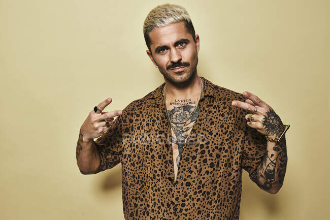 Positive young bearded guy in stylish leopard shirt revealing tattooed torso showing peace gesture while standing against beige background — Stock Photo