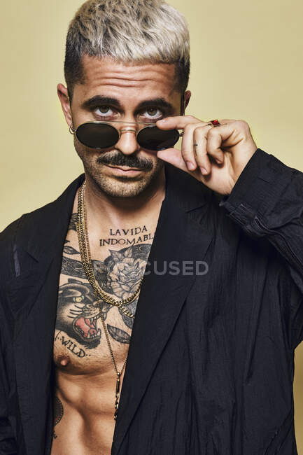 Портрет жорстокого м'язового сексуального чоловіка з татуйованим торсом у чорному пальто та стильними сонцезахисними окулярами та аксесуарами, що стоять на бежевому фоні, дивлячись на камеру — стокове фото