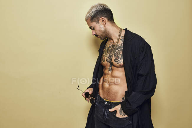 Жестокий мускулистый сексуальный мужчина с татуированным туловищем в черном пальто и модных джинсах со стильными солнцезащитными очками и аксессуарами, стоящими на бежевом фоне, отводя взгляд — стоковое фото