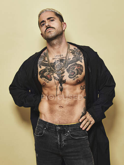 Hombre elegante arrogante seguro con torso musculoso tatuado con abrigo negro y jeans mirando a la cámara sobre fondo beige - foto de stock