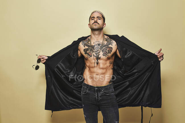 Brutal muscular sexy fit male opening with hands black coat showing up naked tattooed torso while on trendy jeans with stylish sunglasses standing against beige background looking at camera — Stock Photo