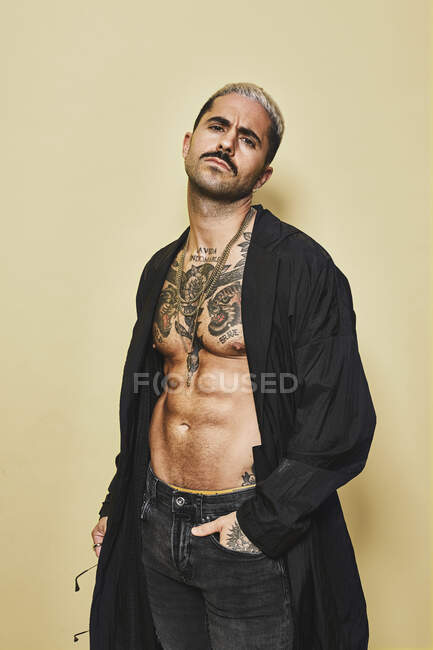 Жорстокий м'язистий сексуальний чоловік з татуйованим торсом у чорному пальто та тенденції та аксесуари, що стоять на бежевому фоні, дивлячись на камеру — стокове фото