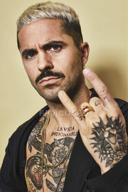 Young provocative male with mustache in black coat over naked tattooed torso showing rock sign gesture while standing against beige background looking at camera — Stock Photo