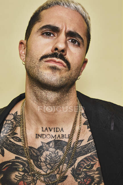 Hombre elegante arrogante confiado con torso musculoso tatuado con abrigo negro mirando a la cámara sobre fondo beige - foto de stock
