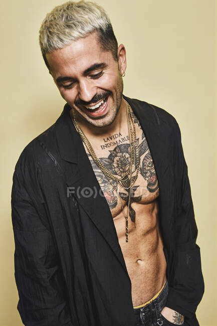 Hombre con estilo alegre y seguro con bigote mostrando su musculoso torso tatuado usando un abrigo negro mirando hacia abajo sobre un fondo beige - foto de stock