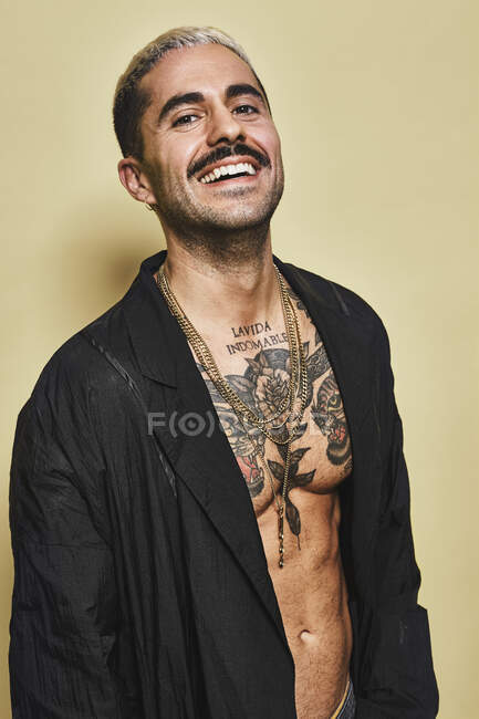 Уверенный в себе веселый стильный мужчина с усами, демонстрирующий свое мускулистое татуированное туловище в черном пальто, смотрящий в камеру на бежевом фоне — стоковое фото