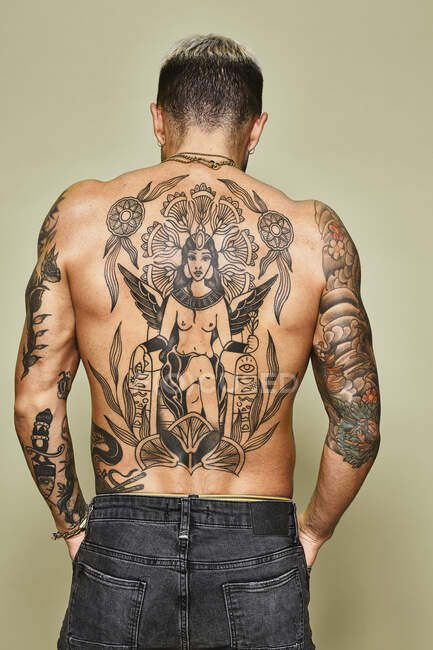Вид ззаду невпізнаваного чоловіка з м'язистим татуйованим тілом у джинсах, що стоять на бежевому фоні — стокове фото