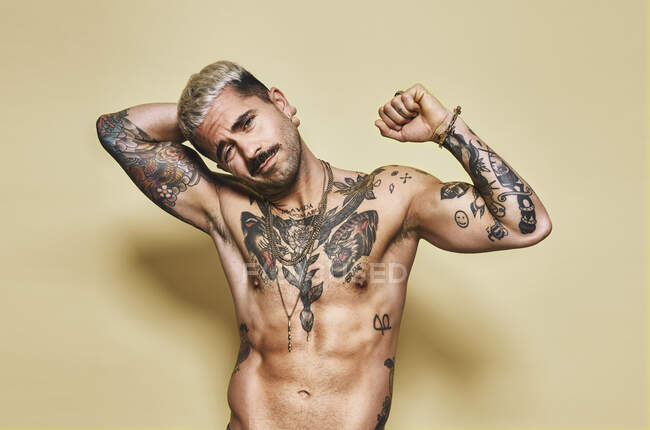 Beau sexy attrayant musculaire mâle avec divers tatouages sur torse nu et les bras en regardant la caméra tout en se tenant debout sur fond beige — Photo de stock