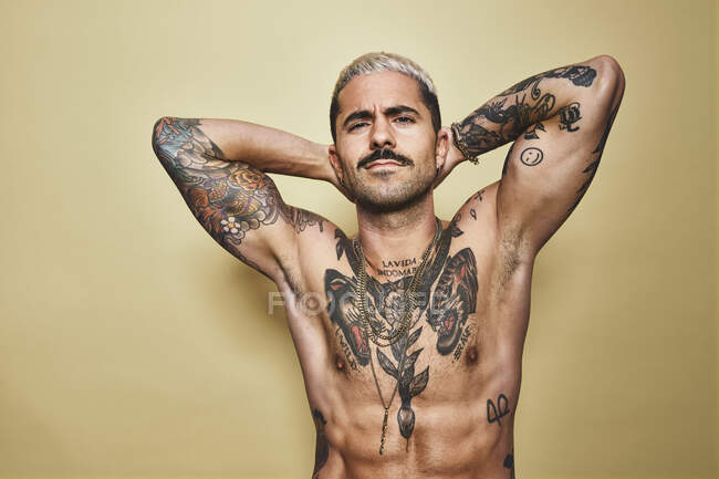 Beau mâle musclé sexy avec divers tatouages sur le torse nu et les bras en regardant la caméra tout en se tenant debout sur fond beige — Photo de stock