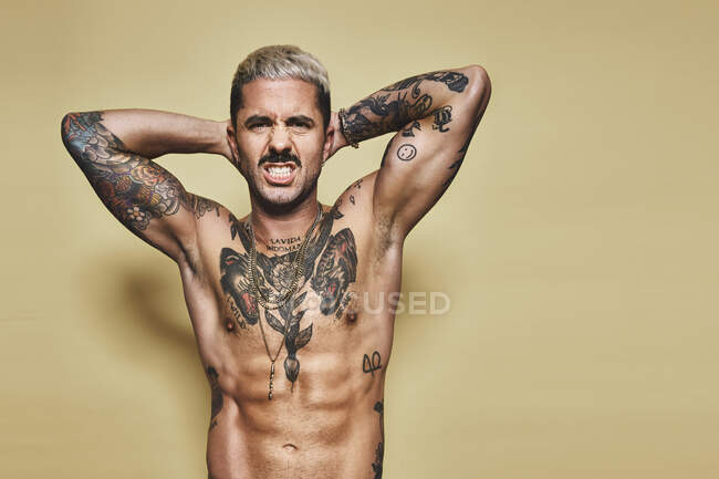 Красивый сексуальный привлекательный мускулистый мужчина с различными татуировками на обнаженном туловище и руках, смотрящий в камеру, корчащий рожи, стоя на бежевом фоне — стоковое фото