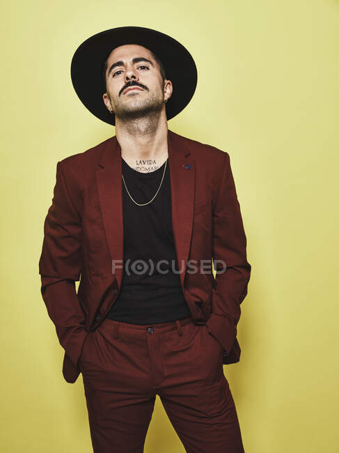 Schöner bärtiger, gut gekleideter Mann im trendigen weinroten Anzug und Hut, der vor gelbem Hintergrund in die Kamera blickt — Stockfoto