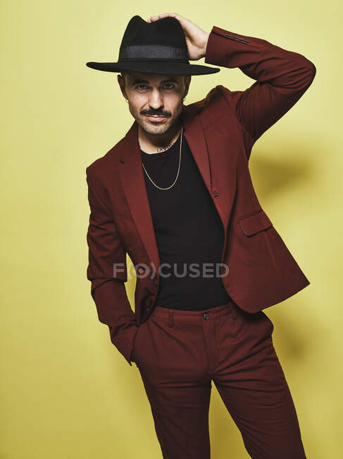 Красивый бородатый мужчина в модном винном костюме и шляпе смотрит в камеру на жёлтом фоне — стоковое фото