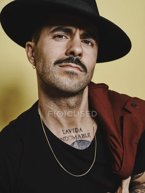 Retrato de hombre guapo bien vestido con bigote en sombrero de moda y traje vinoso que sostiene el abrigo del traje sobre el hombro mirando a la cámara mientras está de pie sobre el fondo amarillo - foto de stock