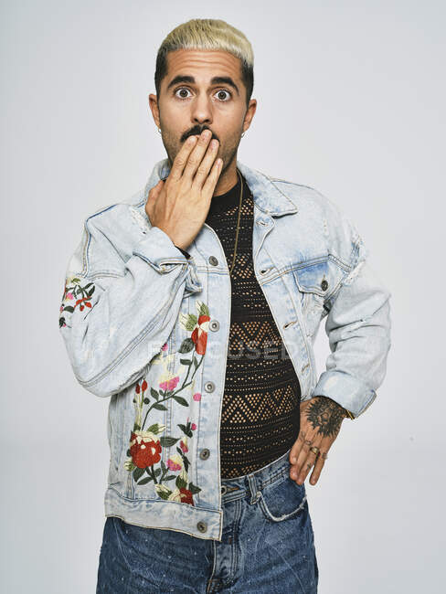 Surprised adult male in stylish denim jacket with floral embroidery covering mouth and looking at camera against gray background — Stock Photo