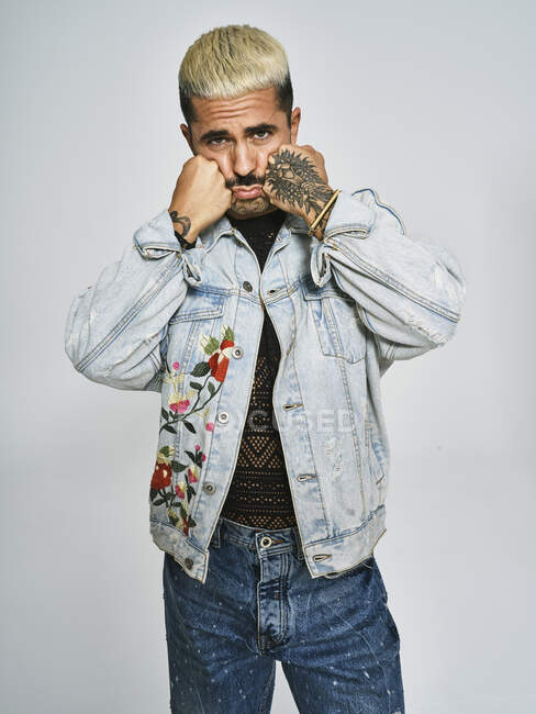 Молодий етнічний чоловік робить похмуре обличчя, дивлячись на камеру в модній джинсовій куртці з квітковим візерунком, стоячи на сірому фоні — стокове фото