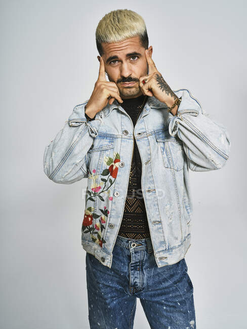 Young ethnic man making grimace doubting face with finger looking at camera wearing trendy denim jacket with floral pattern while standing against gray background — Stock Photo