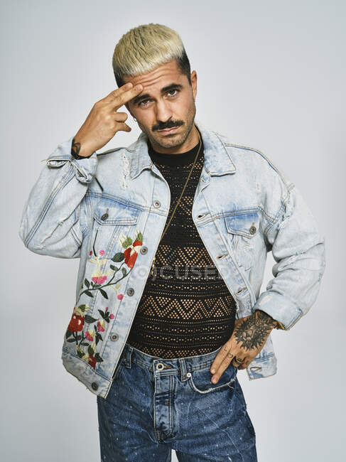 Homem étnico jovem fazendo careta rosto duvidoso com o dedo olhando para a câmera vestindo jaqueta de ganga na moda com padrão floral, enquanto em pé contra fundo cinza — Fotografia de Stock