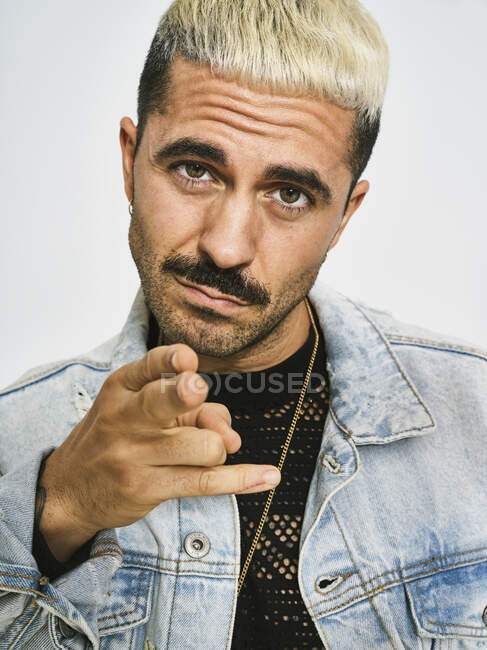 Young ethnic bearded man with trendy hairdo dressed in denim jacket pointing at camera with challenge while standing against gray background — Stock Photo