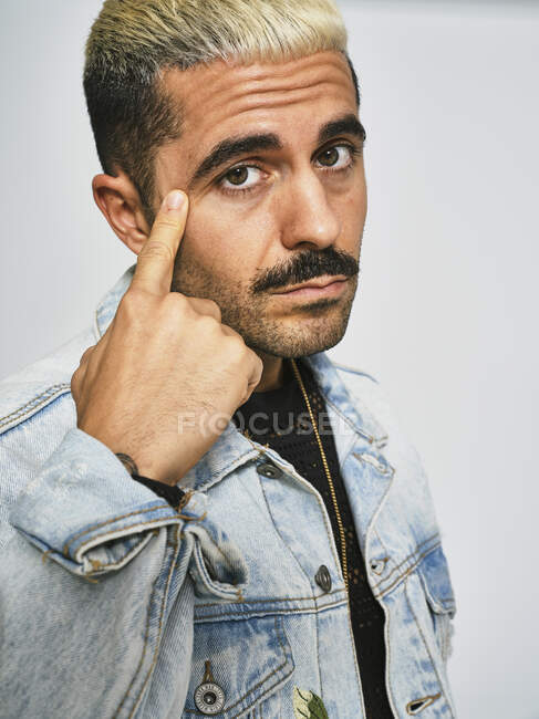 Portrait of young ethnic man making grimace doubting face with finger looking at camera wearing trendy denim jacket with floral pattern while standing against gray background — Stock Photo