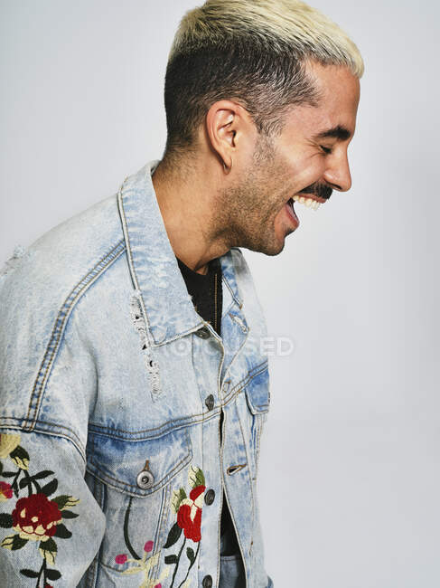 Вид сбоку на молодого стильного парня в модном джинсовом пиджаке, смеющегося стоя на сером фоне — стоковое фото