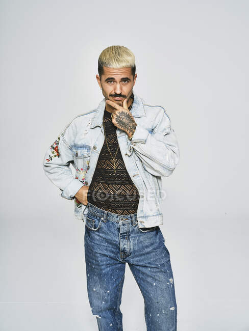 Young ethnic man making grimace doubting face looking at camera wearing trendy denim jacket with floral pattern while standing against gray background — Stock Photo