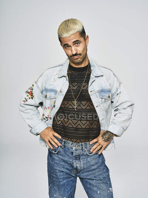 Молодой этнический человек гримасничает, глядя в камеру в модном джинсовом пиджаке с цветочным узором, стоя на сером фоне — стоковое фото