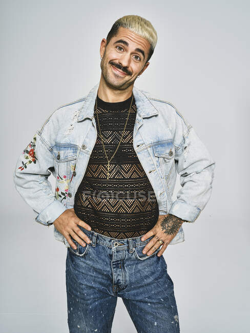Young ethnic man making grimace face looking at camera wearing trendy denim jacket with floral pattern while standing against gray background — Stock Photo