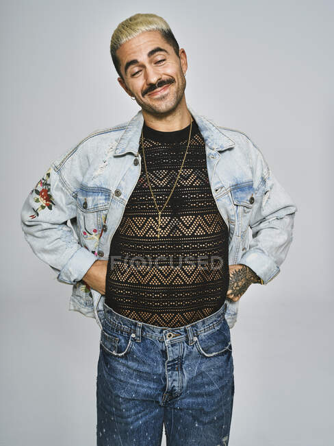 Homem étnico jovem fazendo rosto de careta olhando para a câmera vestindo jaqueta de ganga na moda com padrão floral, enquanto em pé contra fundo cinza — Fotografia de Stock