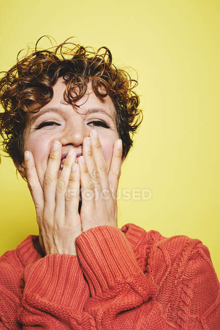 Incroyable femme rieuse aux cheveux bouclés portant un pull tricoté orange couvrant la bouche avec les mains et regardant loin tout en se tenant debout sur fond jaune — Photo de stock