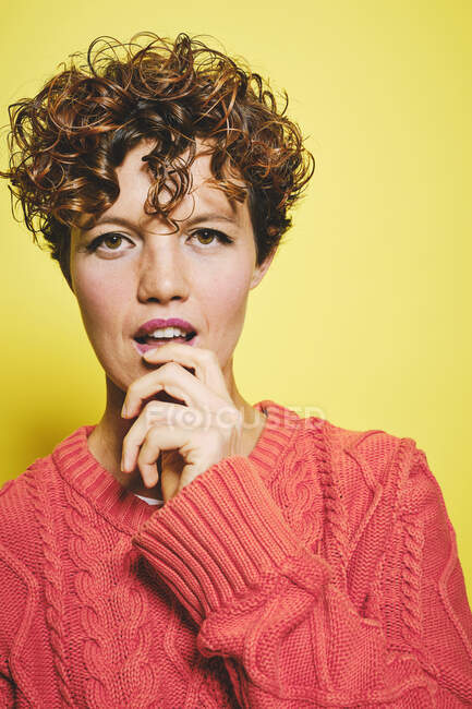 Femme incroyable avec des cheveux bouclés portant un pull tricoté orange avec la main sur la bouche en regardant la caméra tout en se tenant debout sur fond jaune — Photo de stock