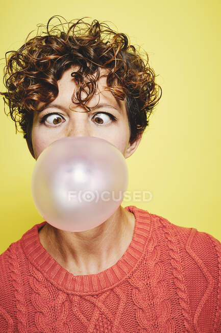 Young curly haired female in orange sweater blowing bubble gum and making funny grimace while standing against yellow background — Stock Photo