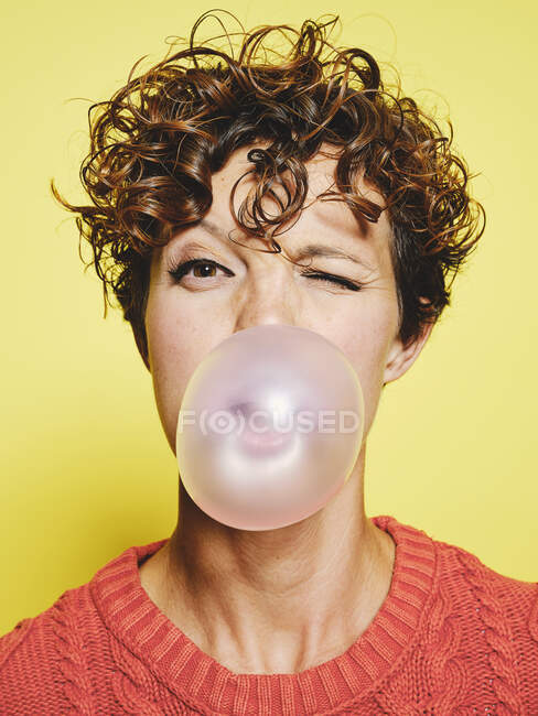 Young curly haired female in orange sweater blowing bubble gum and blinking looking to the camera while standing against yellow background — Stock Photo