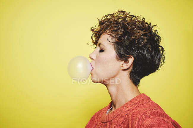 Vue latérale d'une jeune jolie femelle aux cheveux bouclés en chandail orange soufflant de la gomme à bulles debout les yeux fermés sur fond jaune — Photo de stock