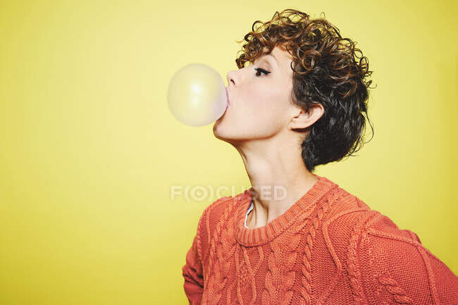 Side view of young pretty curly haired female in orange sweater blowing bubble gum while standing looking away against yellow background — Stock Photo