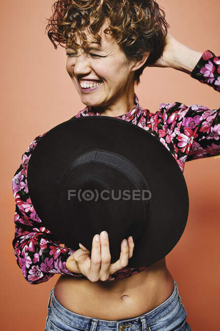 Modelo femenino feliz de moda sosteniendo un elegante sombrero negro en la parte superior de la cosecha de colores con estampado floral de pie sobre fondo naranja con los ojos cerrados - foto de stock