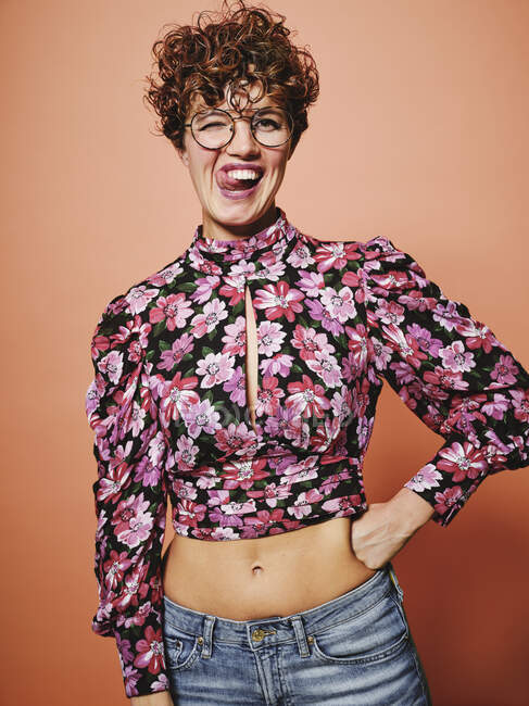 Beautiful curly haired female in trendy eyeglasses and stylish colorful blouse with floral ornament looking at camera and making funny grimace with tongue out standing against pink background — Stock Photo