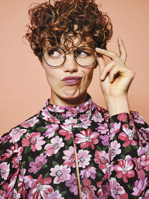 Задумчивая сомнительная красивая кудрявая женщина в модных очках и стильной красочной блузке с цветочным орнаментом, смотрящей в сторону на розовом фоне — стоковое фото
