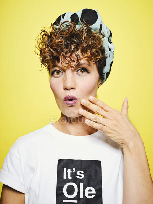 Portrait of adult female in stylish outfit keeping hand on cheeks and looking at camera in shock against yellow background — Stock Photo
