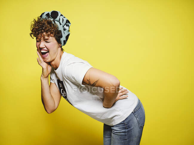 Adult female in trendy casual outfit with head wrap with closed eyes and laughing out loud at funny joke while standing against yellow background — Stock Photo