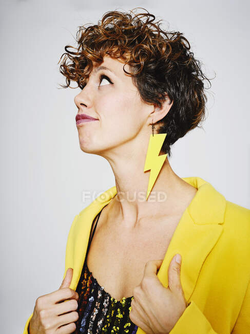 Портрет задумчивой женщины с молниеносной сережкой и блестками сверху, регулирующий стильное желтое пальто на сером фоне, глядя вверх — стоковое фото