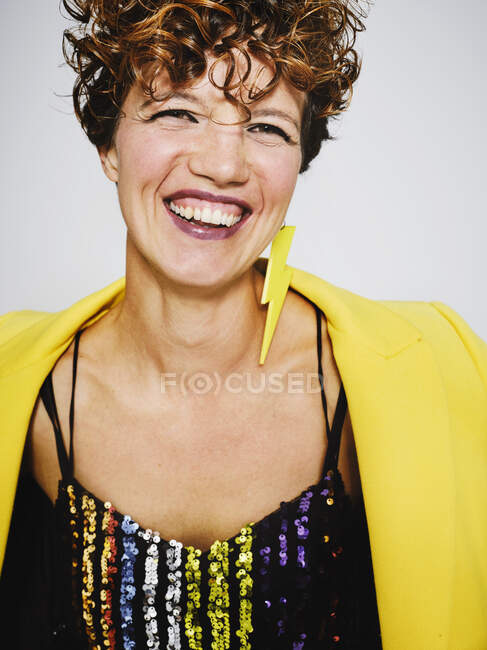 Portrait of cheerful woman with sequin top and lightning earring and stylish yellow coat smiling to the camera while standing against gray background — Stock Photo
