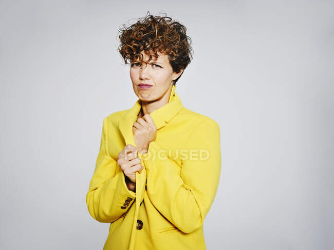 Porträt einer kalten Frau mit Blitz-Ohrring, der einen eleganten gelben Mantel vor grauem Hintergrund vor die Kamera hält — Stockfoto